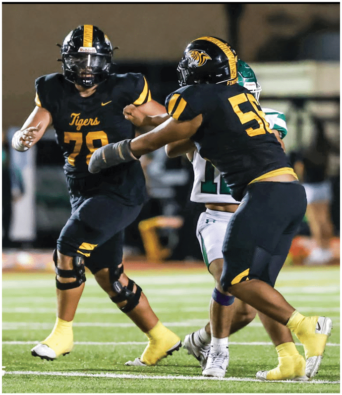 Two football players in black and yellow uniforms are actively engaged with an opposing player in white and green on a field during a game. Description generated by AI