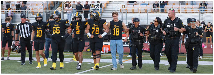 A group of people wearing football uniforms and helmets stand together on a sports field with bleachers and spectators in the background. Description generated by AI