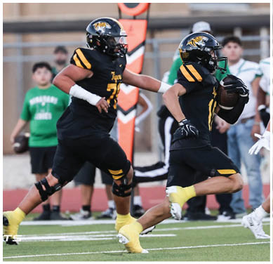 Two people in black and yellow football uniforms are running on a field, with one holding a football and the other following closely behind. Description generated by AI