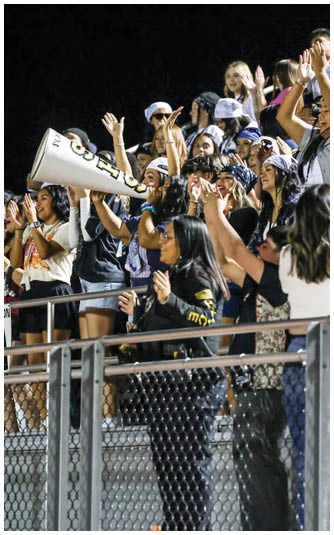 A group of enthusiastic people standing in bleachers at night, cheering and raising their hands, with one person holding a large megaphone. Description generated by AI
