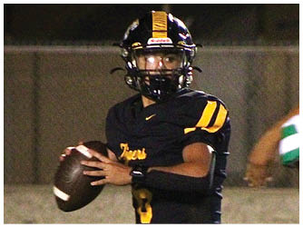 A person in a black football uniform with yellow stripes is holding a football and preparing to throw it while another person in a white and green uniform approaches. Description generated by AI