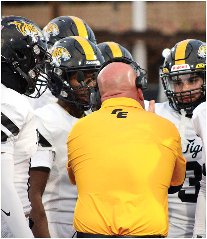 A group of people wearing football helmets and white jerseys are gathered around a person in a yellow shirt with a headset, likely during a game or practice. Description generated by AI