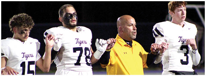 Four people stand holding hands, three wearing white football uniforms with “Tigers" on the front and one in a yellow jacket, appearing to be on a sports field at night. Description generated by AI