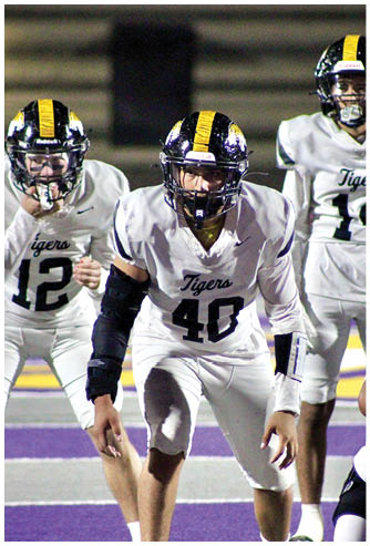Three people in white football uniforms with black and yellow helmets are positioned on a field, appearing ready for action. Description generated by AI