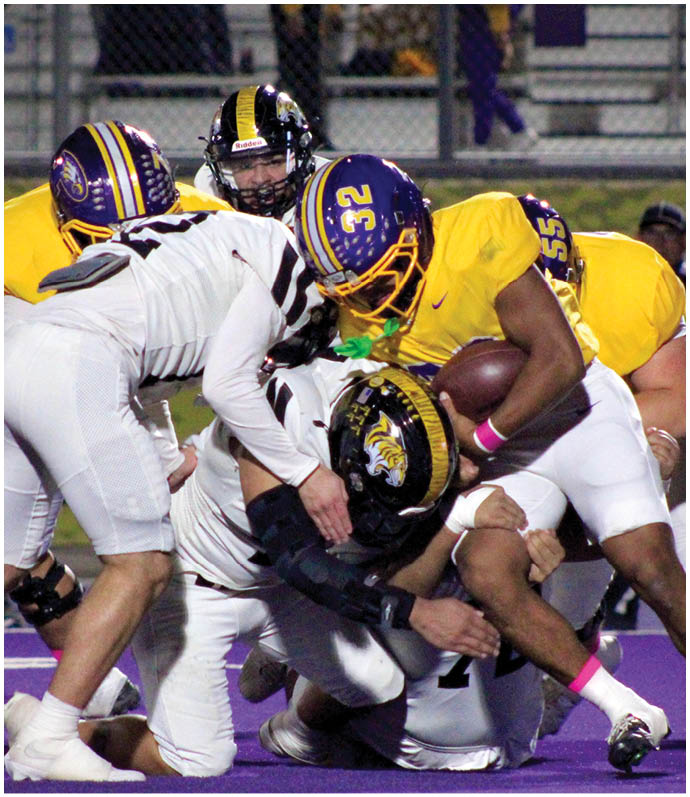 Several people in football uniforms are engaged in a tackle on a purple field during a game. Description generated by AI