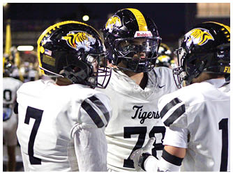 Three people wearing white football uniforms with black and yellow helmets featuring a tiger logo are standing together on a field at night. Description generated by AI