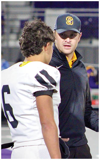 A person wearing a yellow Under Armour polo shirt looks to the side with a neutral expression while another person with curly hair is partially visible. Description generated by AI