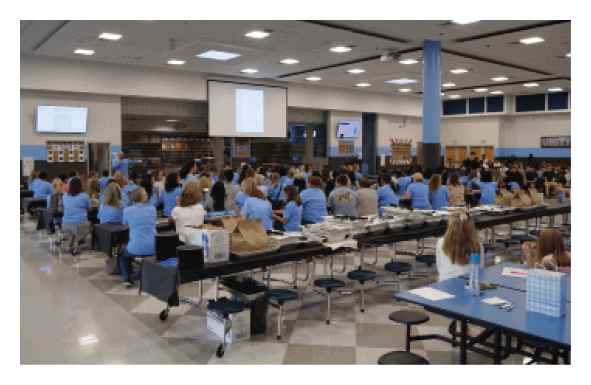 A large group of people are sitting in a room, with some standing and others sitting at tables. The room is filled with chairs and dining tables, and there are TVs on the walls. The people are engaged in a presentation or meeting, with some of them holding bags. AI generated content