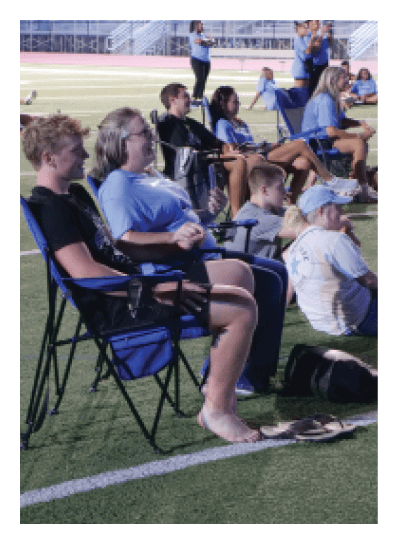 A group of people are sitting on a field, some on chairs and others on the ground. They are watching something, possibly a game or an event. AI generated content