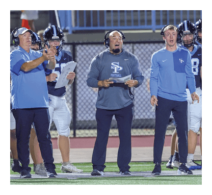 A group of men wearing blue shirts and white pants are standing on a field. They are wearing headsets and appear to be listening to a coach. AI generated content