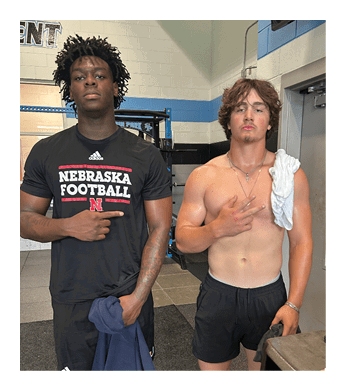 Two men standing in a gym, one shirtless and the other wearing a black shirt with the word “nebraska" on it. AI generated content