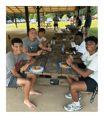 Four young men are sitting at a picnic table, enjoying a meal together. They are wearing shorts and are in a relaxed setting. There are several cars parked in the background, and a bench is visible in the foreground. The atmosphere appears to be casual and friendly. AI generated content