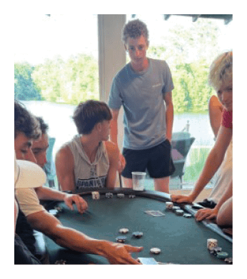 A group of people are playing poker at a table, with some of them sitting on chairs and others standing. There are several cups and a bowl on the table, along with a variety of cards. The players are engaged in the game, with some of them holding cards in their hands. AI generated content