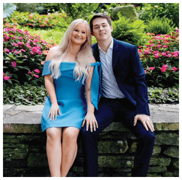A man and woman are sitting on a stone wall, posing for a picture. The woman is wearing a blue dress and the man is wearing a suit. AI generated content