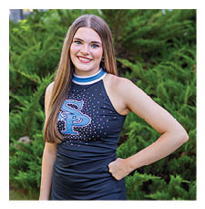A young woman wearing a cheerleading outfit and a blue skirt stands in front of a bush. AI generated content