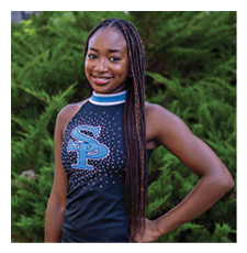A young woman wearing a black and blue cheerleading outfit stands in front of a bush. AI generated content