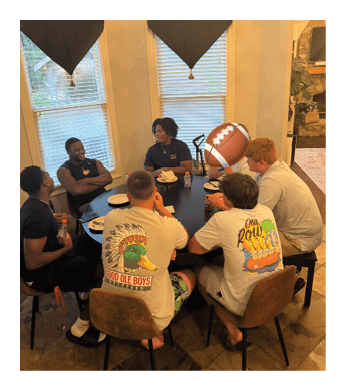 A group of people are sitting around a table, eating and drinking. There are four people on one side of the table and three on the other. A football is present on the table, and a bottle is also visible. The people appear to be enjoying their time together. AI generated content