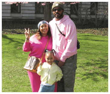 A man and woman pose with a child in a park. The woman is wearing pink and holding a handbag. The man is wearing a pink hoodie. AI generated content