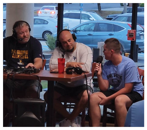 Three men are sitting around a table, engaged in a conversation. One of them is wearing a headset, possibly a radio host. They are surrounded by chairs and a dining table. In the background, there are several cars parked outside. AI generated content