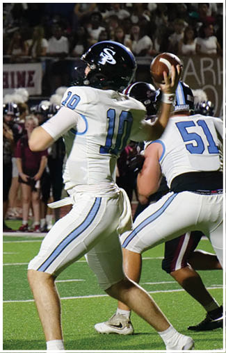 A football game is taking place with a player in a white jersey getting ready to throw the ball. The player is wearing a blue and white uniform and is surrounded by other players on the field. The crowd is watching the game intently, and there are several people in the background. AI generated content