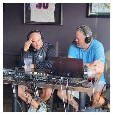 Two men are sitting at a table with a laptop and a microphone. One of the men is wearing headphones. AI generated content