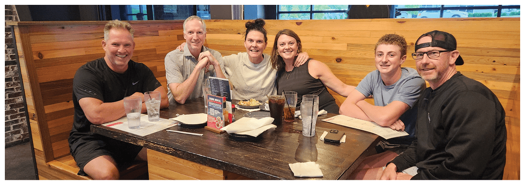 A group of people are sitting around a wooden table in a restaurant, posing for a picture. They are smiling and enjoying their time together. There are several cups, a cell phone, and a book on the table. AI generated content