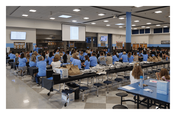 A large group of people are sitting in a room, with some standing and others sitting at tables. The room is filled with chairs and dining tables, and there are TVs on the walls. The people are engaged in a presentation or meeting, with some of them holding bags. AI generated content