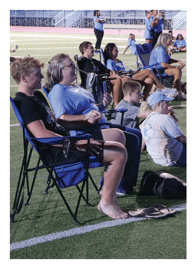 A group of people are sitting on a field, some on chairs and others on the ground. They are watching something, possibly a game or an event. AI generated content