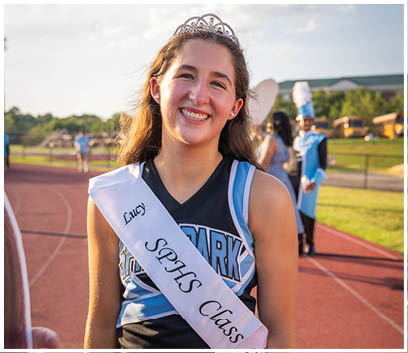 A girl wearing a blue and white dress and a sash that says “Sparks Class 9" is smiling. AI generated content