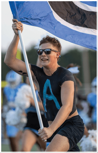 A man in a black shirt and white shorts holds a flag while a cheerleader in a blue and white uniform stands behind him. AI generated content