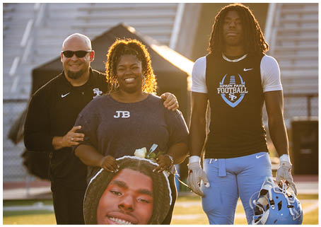 A man and woman are standing on a field with a football player in the background. The woman is holding a football player's head on a board. AI generated content