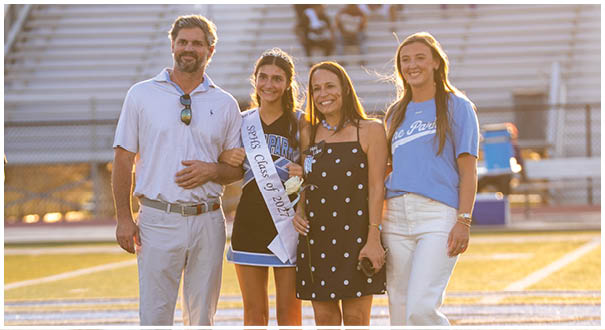 A group of people, including a man, woman, and two girls, are standing on a field. The woman is holding a sash, and the girls are wearing dresses. They are posing for a picture. AI generated content