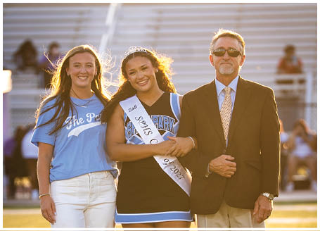 Three people are standing on a field, two women and a man. The women are wearing blue shirts and white pants, while the man is wearing a suit. The man is holding a microphone. AI generated content
