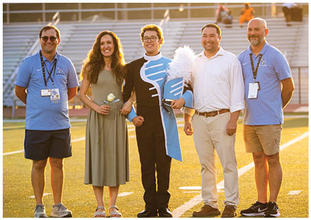 Four people are standing on a field, posing for a picture. Two men and two women are standing together, with one of the women holding a bouquet. The group appears to be a brass band, as one of the men is holding a trumpet. AI generated content