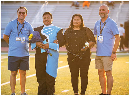 Four people are standing on a field, posing for a picture. Two men and two women are standing together, with the men on the left and the women on the right. The men are wearing ties, and the women are holding flowers. AI generated content