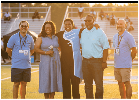 Four people are standing on a field, posing for a picture. Two men and two women are standing together, with one of the women holding a bouquet of flowers. The group appears to be a family or friends, enjoying a special moment together. AI generated content
