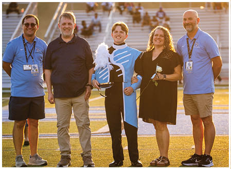 Four people are standing on a field, posing for a picture. Two men and two women are standing together, with one of the men holding a flag. The group appears to be a band, as they are all wearing uniforms. AI generated content