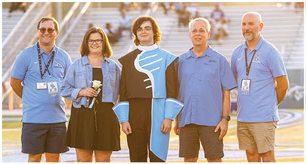 Four people are standing on a field, posing for a photo. A man in a blue and black costume is standing between two men and a woman. The woman is wearing a blue dress. AI generated content