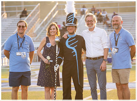 Four people pose for a photo on a football field. A man in a drum major uniform stands between two men and a woman. The woman is holding a bouquet of flowers. AI generated content