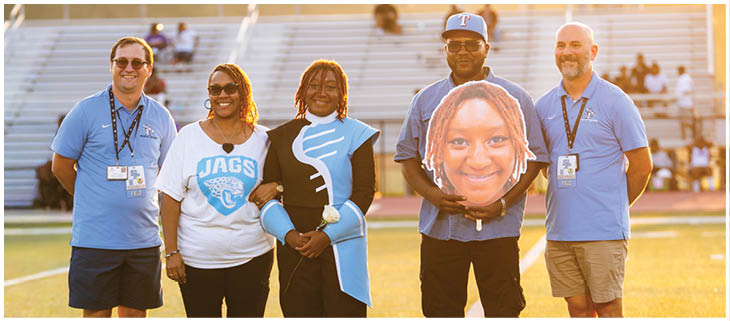 Four people pose for a picture on a football field. They are wearing blue shirts and holding a football shaped like a head. AI generated content