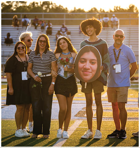 A group of people are standing on a field, posing for a photo. One of the individuals is holding a cutout of a man's face. AI generated content