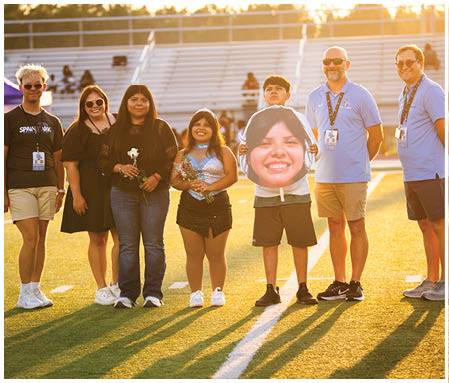 A group of people are standing on a field, posing for a photo. They are holding flowers and a cutout of a smiling face. AI generated content