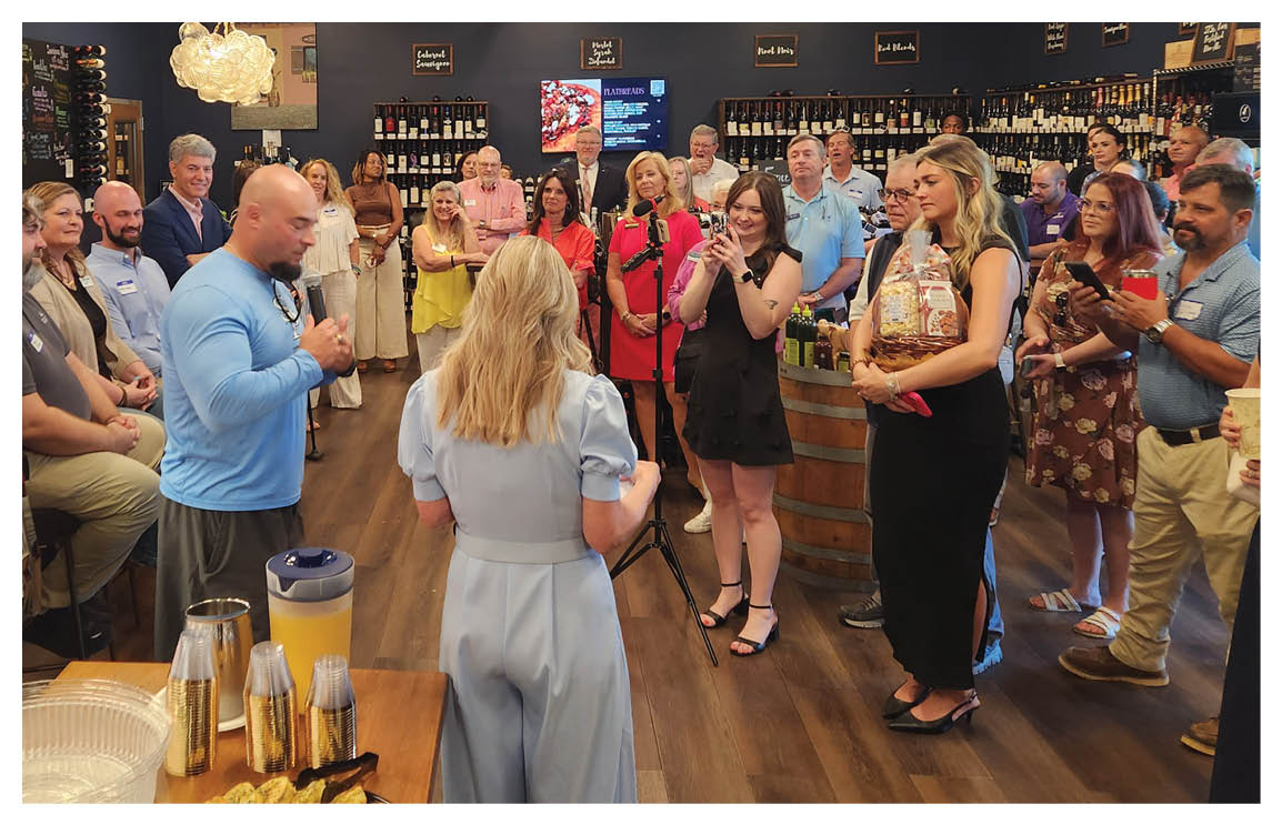 A group of people are standing around a table with food and drinks. A man is holding a camera, possibly taking pictures of the event. The people are engaged in conversation and enjoying the gathering. AI generated content