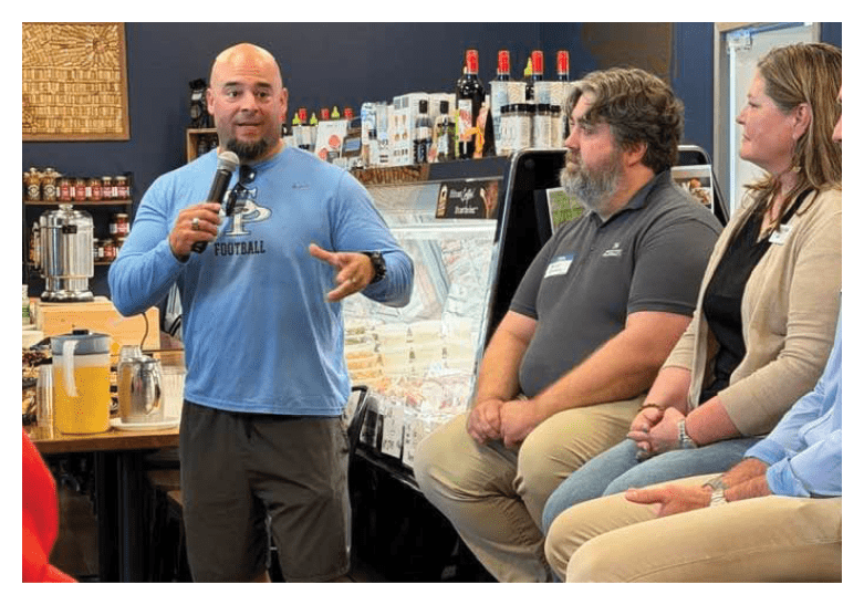 A man in a blue shirt is speaking into a microphone while three people listen. The man is standing in front of a table with a bottle on it. AI generated content