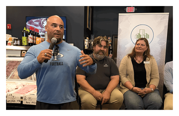 A man in a blue shirt stands in front of a microphone, speaking to a group of people. The group consists of three people, with one person sitting on a chair and the other two standing. The room has a dining table and a refrigerator. There are several bottles and a cup placed around the room. AI generated content