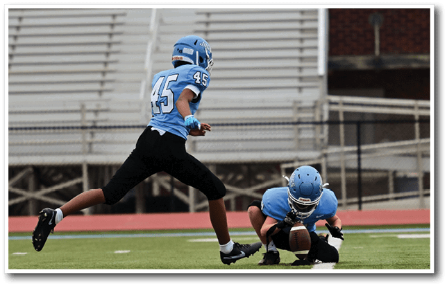 Two football players on a field, one player is about to kick the ball while the other player is kneeling down. AI generated content