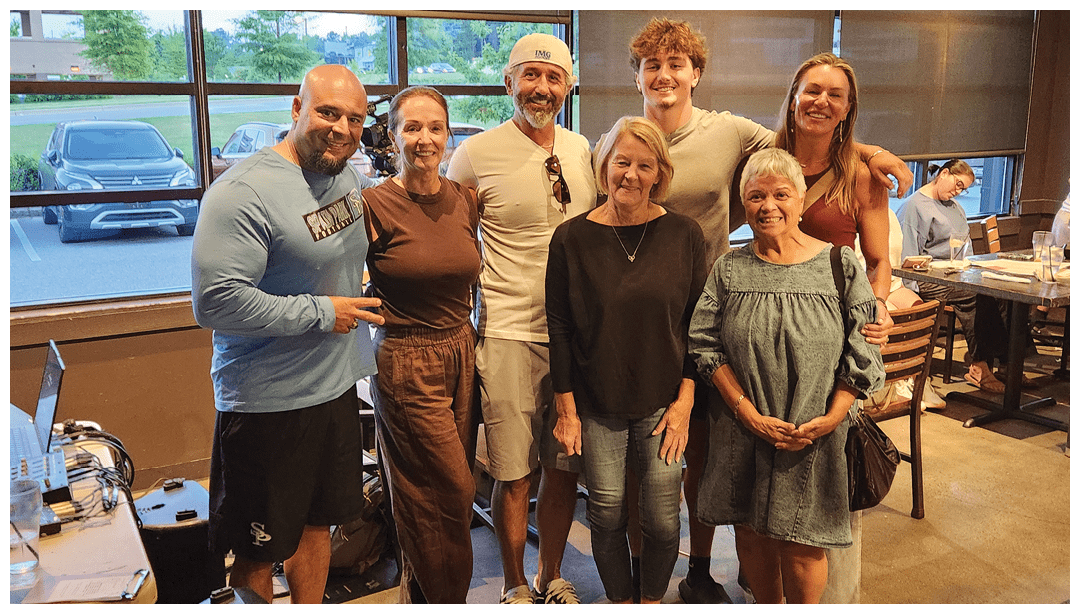 Seven adults stand closely together smiling inside a casual dining setting with large windows showing parked cars outside. Description generated by AI