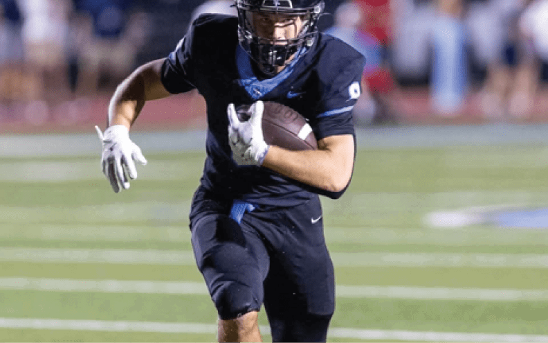Three people in dark football uniforms are actively playing on a field with one person running while holding the ball. Description generated by AI