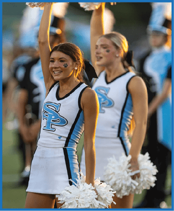 A person in a cheerleading uniform with “SP" on the front is smiling and holding pom-poms up during a performance on a sports field with others in similar uniforms in the background. Description generated by AI