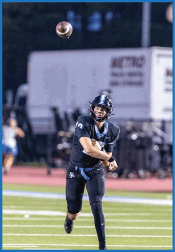 A person in a dark football uniform and helmet is throwing a football on a green field with a blurred background of people and equipment. Description generated by AI